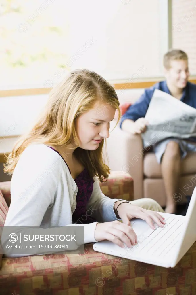 Teenage girl working on a laptop and a teenage boy reading a newspaper beside her