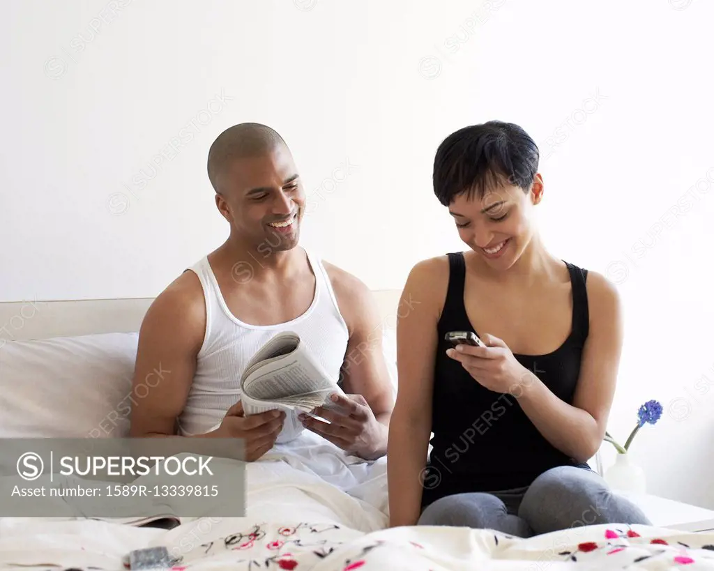 Couple relaxing in bed with newspaper and cell phone