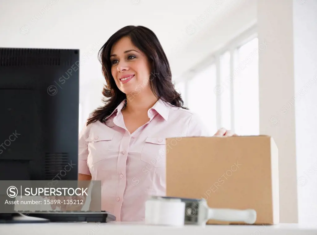 Smiling Hispanic woman with cardboard box using computer