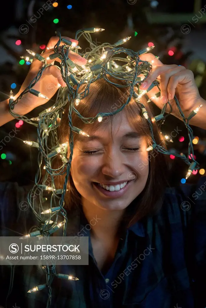 Mixed Race woman holding string lights on head near Christmas tree