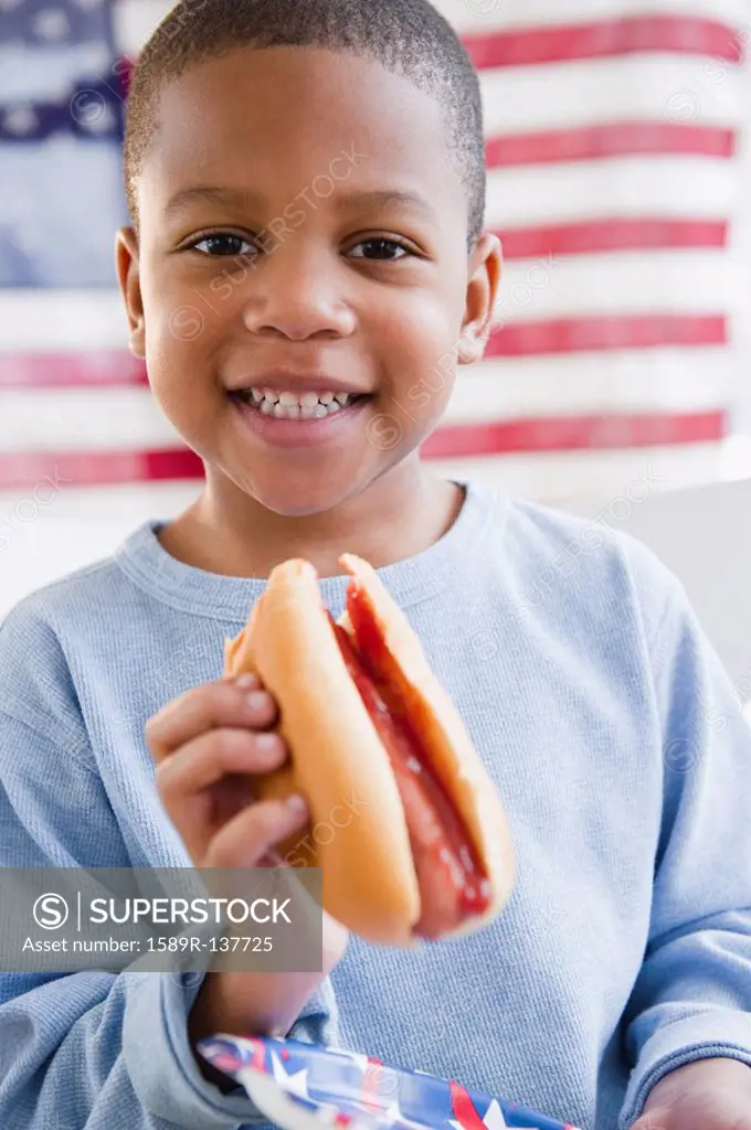 African American boy eating hot dog in front of American flag