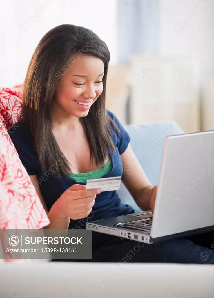Smiling mixed race teenage girl holding credit card and laptop on sofa
