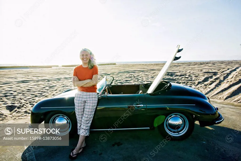 Older Caucasian woman leaning on convertible car with surfboard on beach