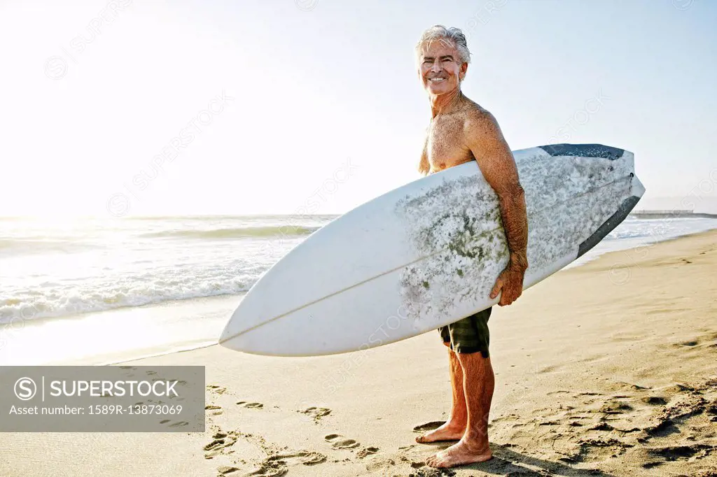 Older Caucasian man standing on beach carrying surfboard