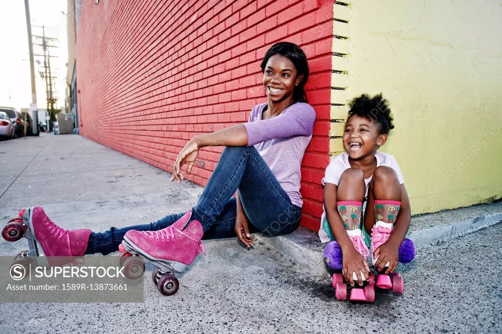 Black mother and daughter wearing roller skates sitting on sidewalk