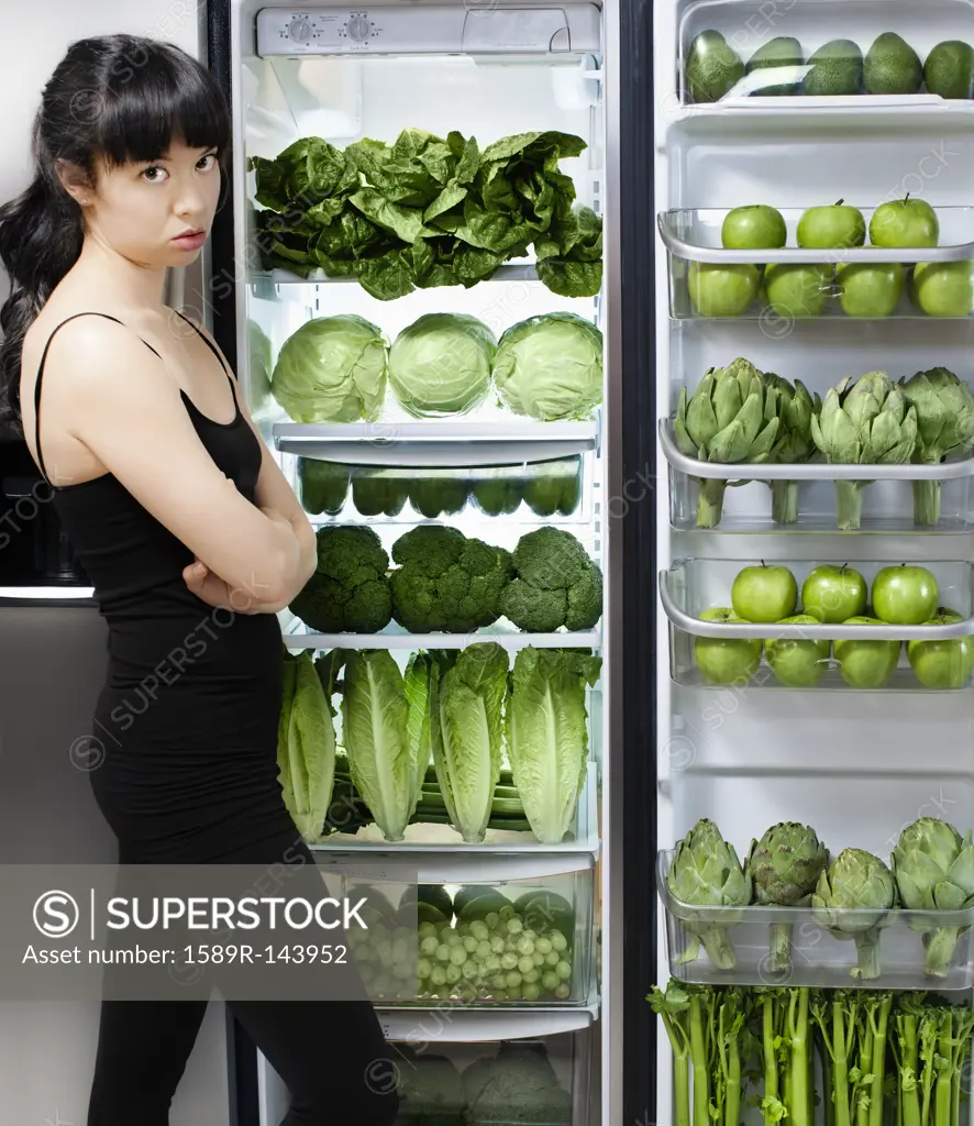 Unhappy mixed race woman looking at green vegetables in refrigerator