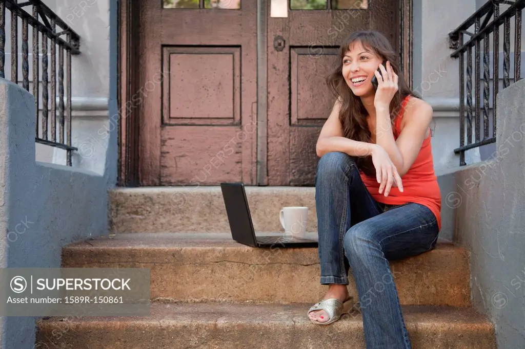 Mixed race woman sitting on front stoop with cell phone and laptop