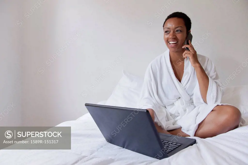 African American woman using laptop and cell phone in bed
