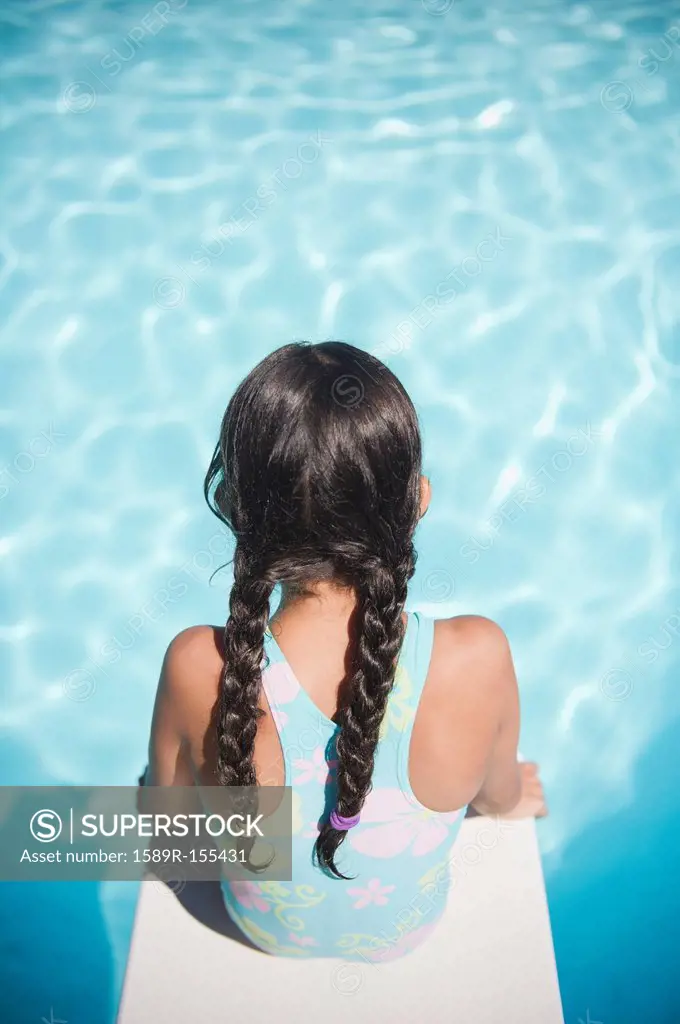 Hispanic girl sitting on diving board over swimming pool