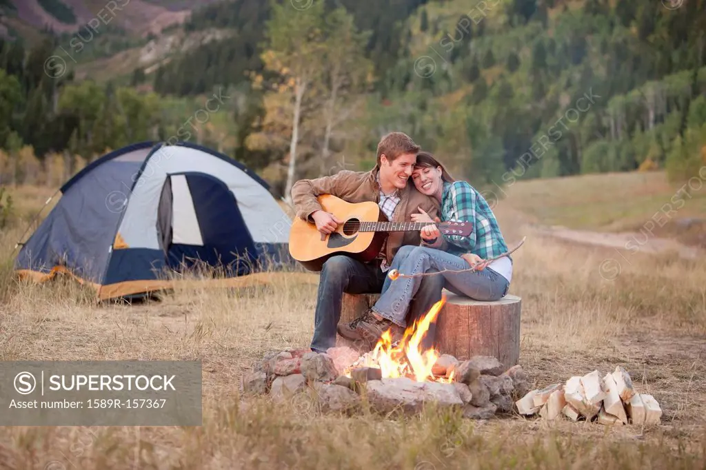 Caucasian man playing guitar for wife near campfire