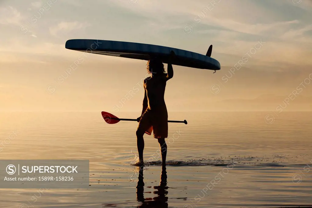 Caucasian man walking in water carrying paddle board