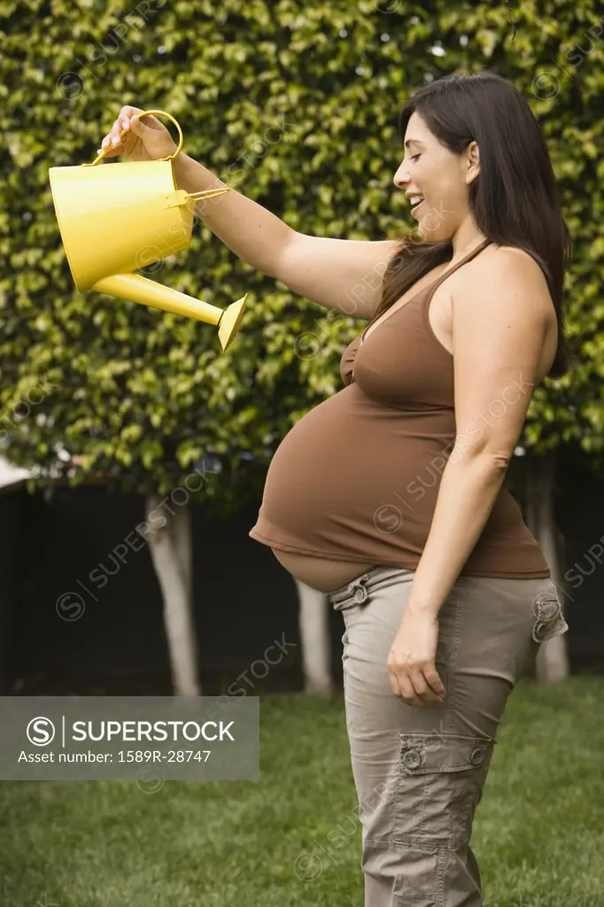 Pregnant Hispanic woman holding watering can over belly outdoors