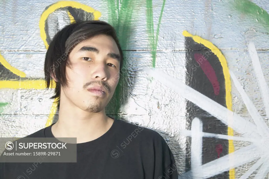 Young Asian man leaning against graffitied wall