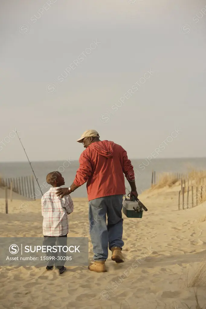 African father and son walking on beach with fishing gear - SuperStock