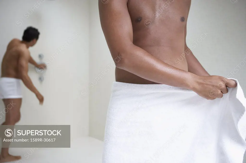 Man wrapping towel around waist while man in underwear tests shower in background