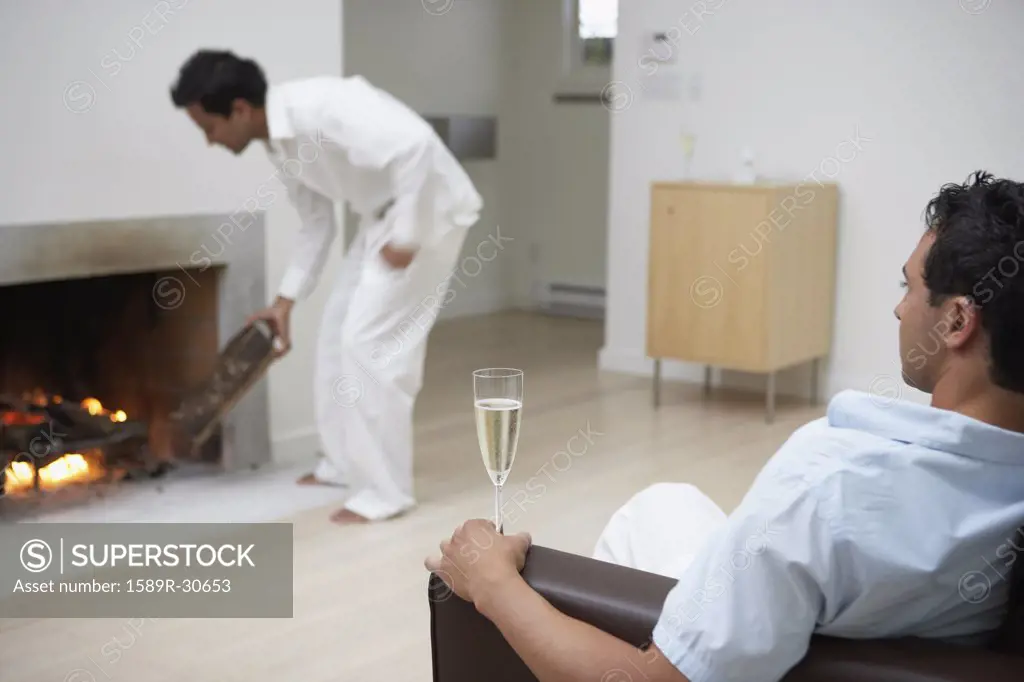 Man on sofa with champagne while male friend puts wood in fireplace