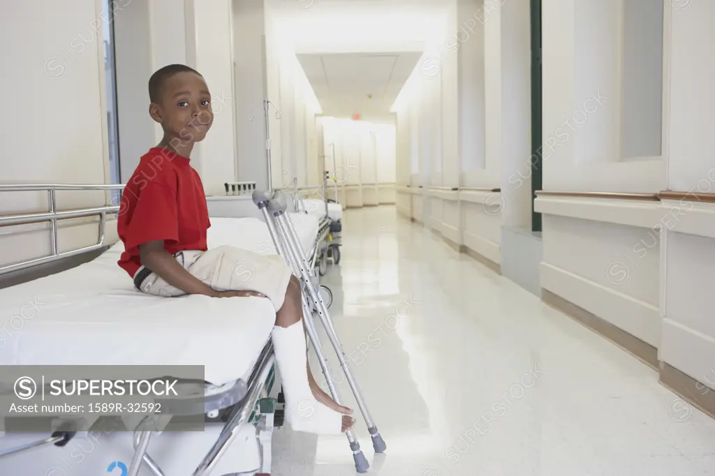 African boy with broken leg and crutches sitting on bed in hospital corridor
