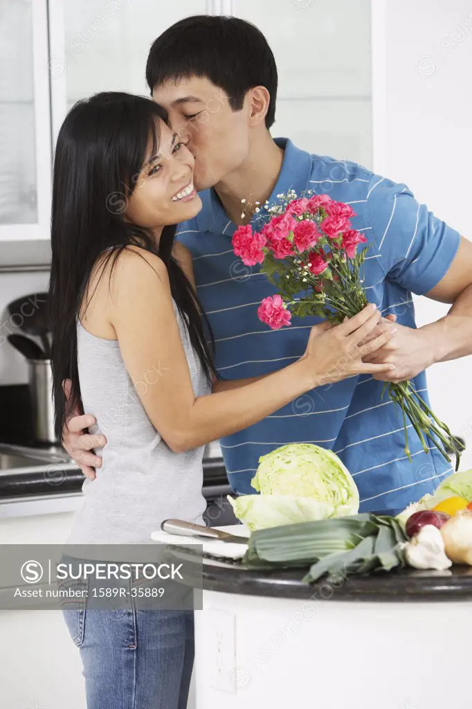 Asian man kissing girlfriend and giving her flowers in kitchen