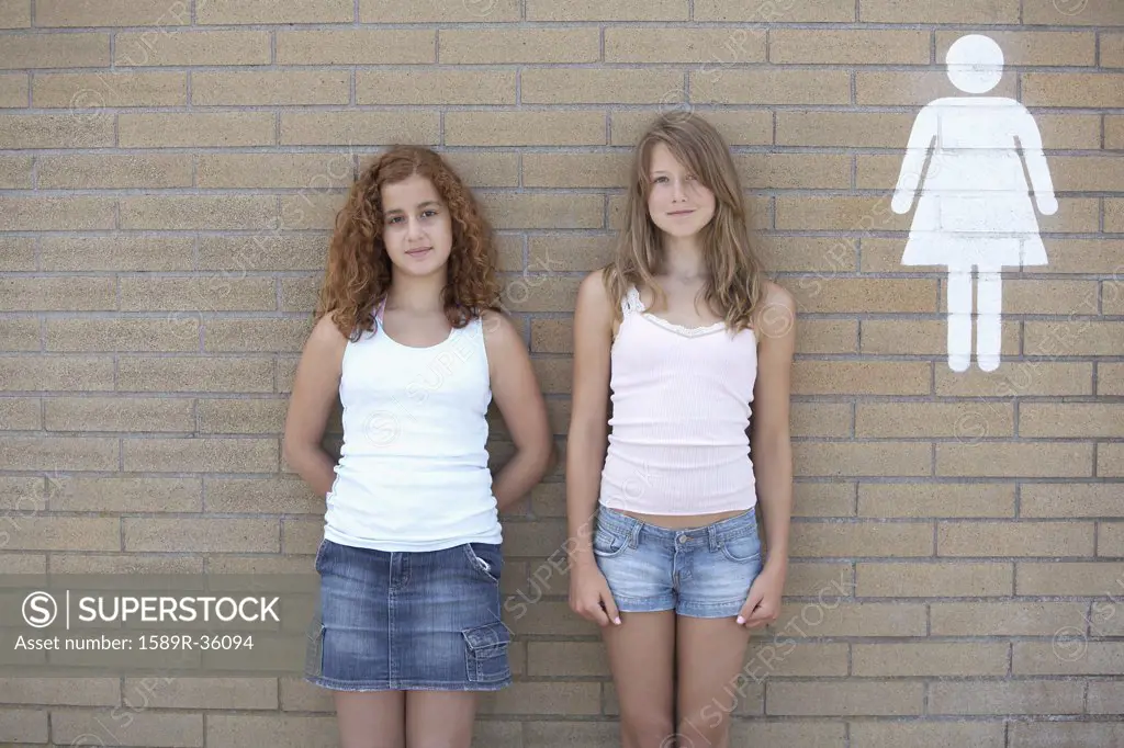 Two teenage girls leaning against wall next to restroom sign