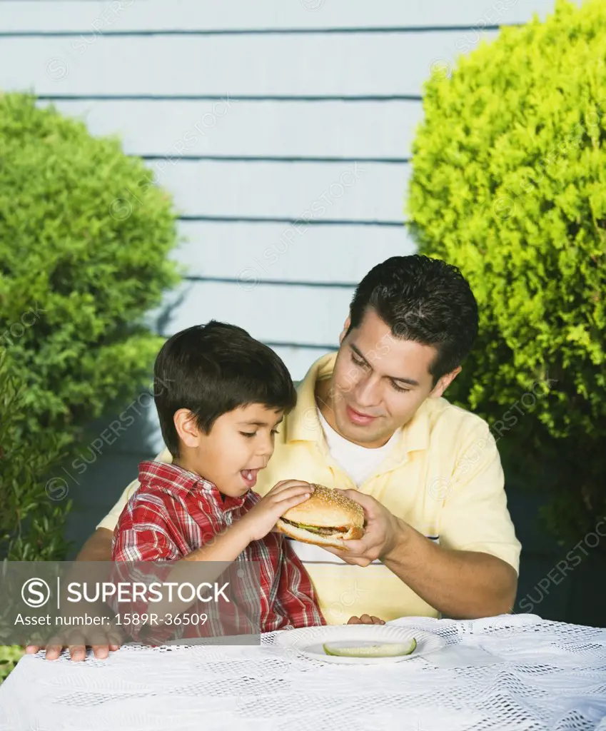 Hispanic father helping young son eat hamburger