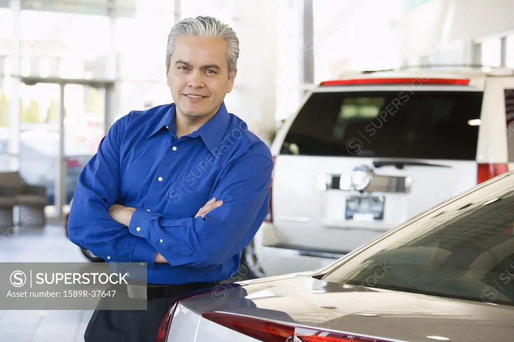 Hispanic car salesman leaning on new car