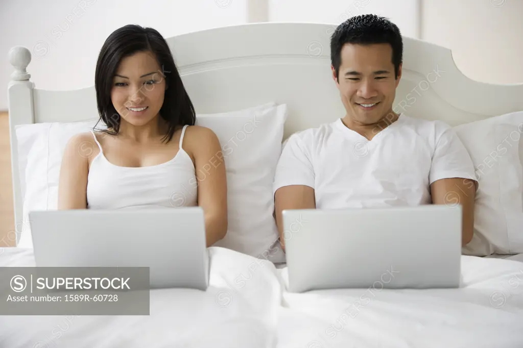 Asian couple typing on laptops in bed