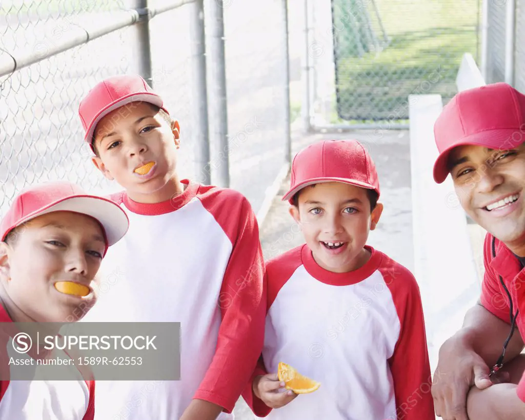 Multi-ethnic boys in baseball uniforms and coach in dugout eating oranges