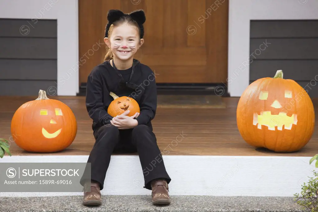 Mixed race young girl in cat costume holding Halloween pumpkin