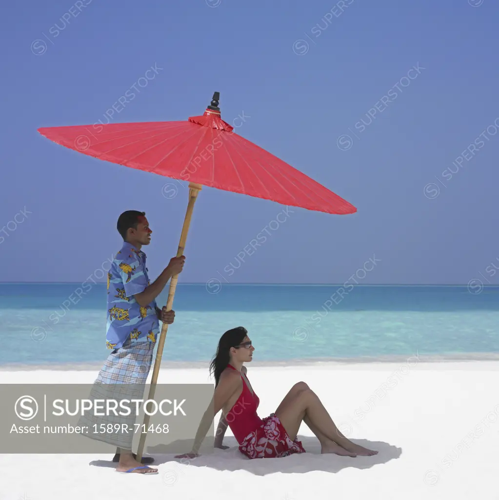 Maldivian man holding beach umbrella over woman