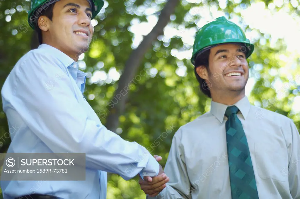 Hispanic businessmen in green hard hats shaking hands outdoors