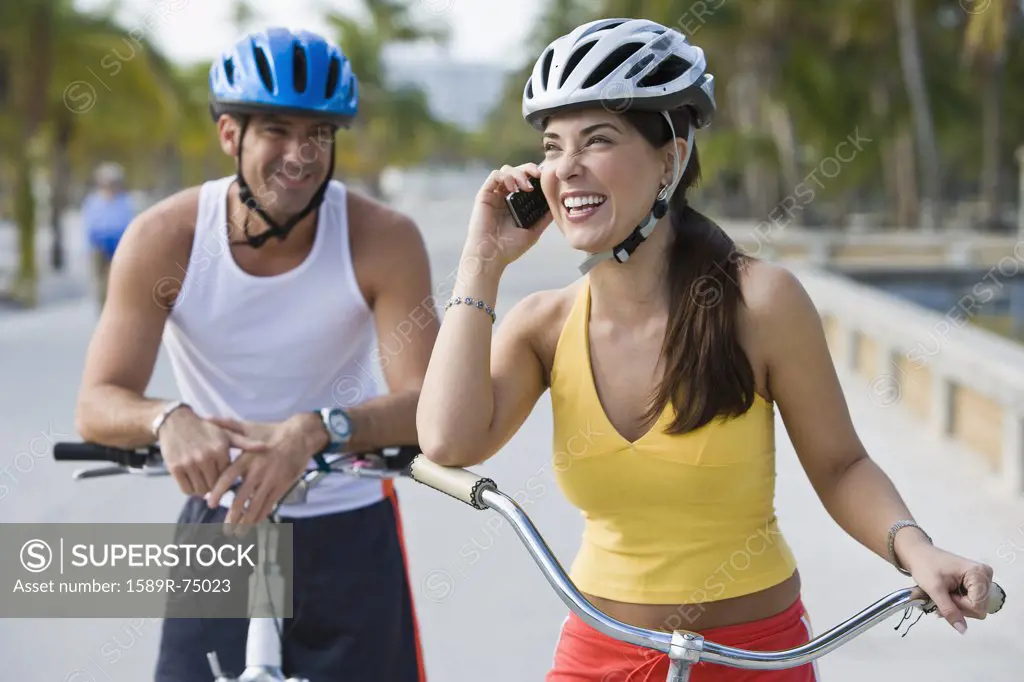 Hispanic woman on bicycle talking on cell phone while boyfriend waits