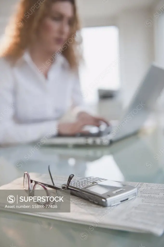 Hispanic woman using laptop near newspaper, cell phone and eyeglasses