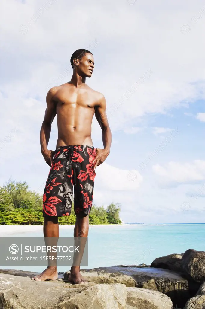 African man in swim trunks standing on rocks by ocean