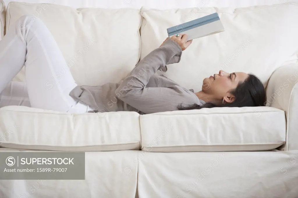 Hispanic woman reading book on sofa
