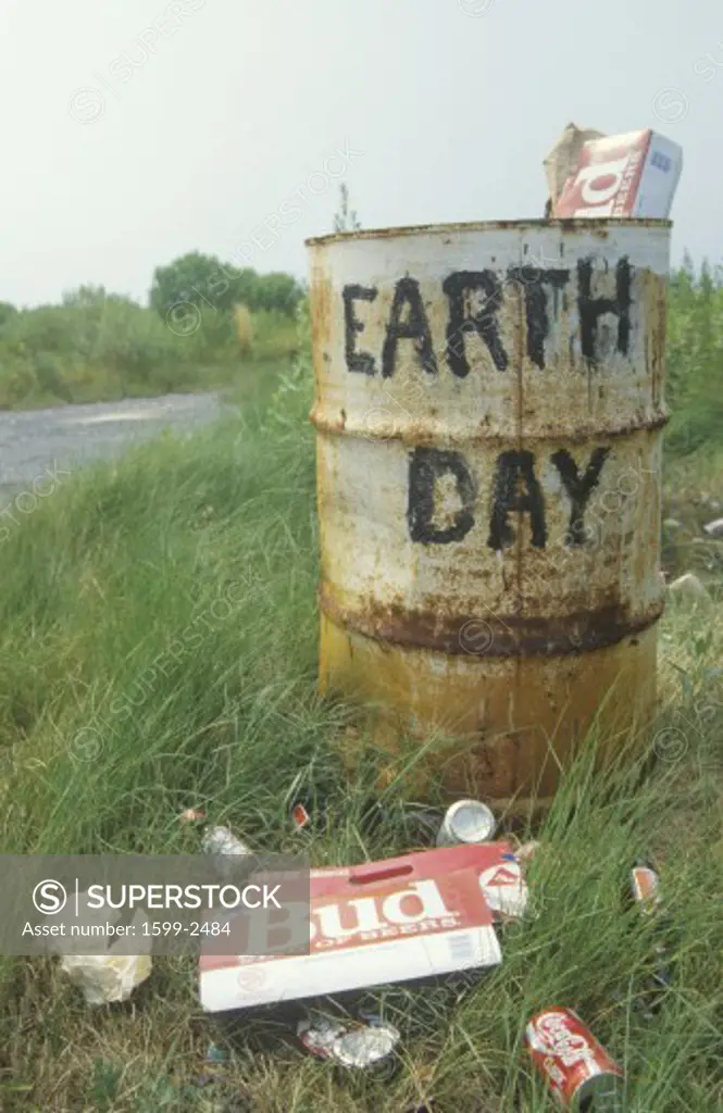 Cardboard beer cartons on the ground next to a trash can with the words Earth Day” painted on its side 