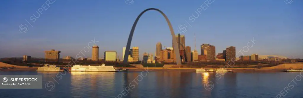 This is the skyline at sunrise. It is situated along the Mississippi River. There are riverboats on the water with the St. Louis Arch in clear view.