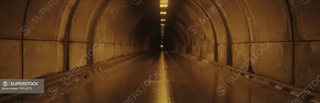 This is the interior of a tunnel. There is an orange glow from lights on the surrounding walls with a small light at the end of the tunnel. There is a road leading through the tunnel.