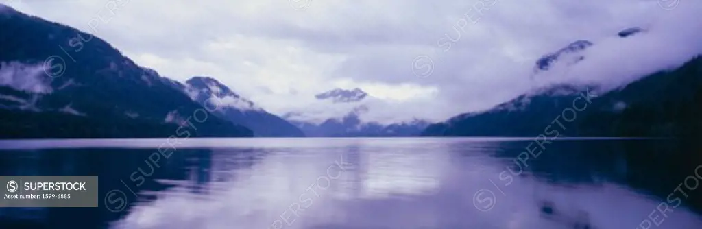 This is an image of the Olympic Peninsula with Crescent Lake in the foreground. The mountains in the background are covered with low altitude clouds. There is a reflection in the lake of the clouds and mountains. The sky is overcast with clouds.