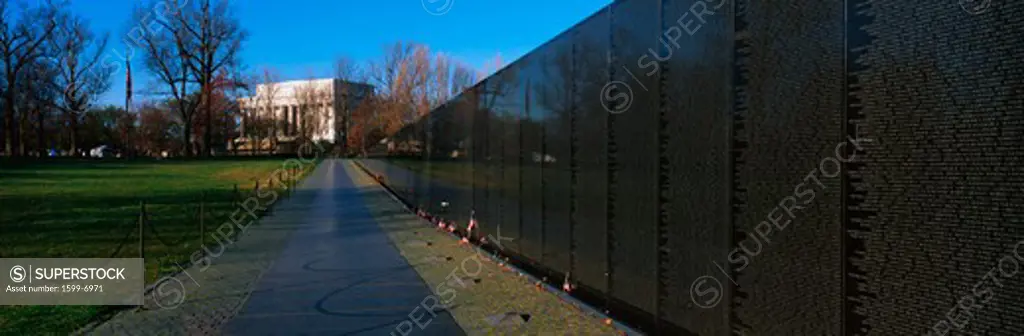 This is the Vietnam Veterans Memorial known as the Wall. A sidewalk leads to the Lincoln Memorial. there are a few small American flags placed up against the bottom of the wall with small flowers. On the other side is a small chain held up by small posts lining the opposite side of the sidewalk.