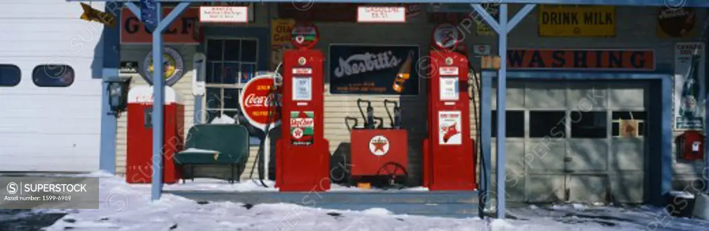 This is a vintage gas station with old style pumps that are red. It is a piece of Americana. It shows winter in New England. There is an old-fashioned Coke sign with other vintage signs. There is snow on the ground with a small bench and a garage door at both ends of the gas station.