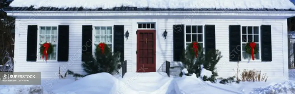 This is a nicer looking home in a winter setting. The house is surrounded by snow. The house has black shutters and a red front door. There are Christmas wreaths in each window. This shows winter in New England.