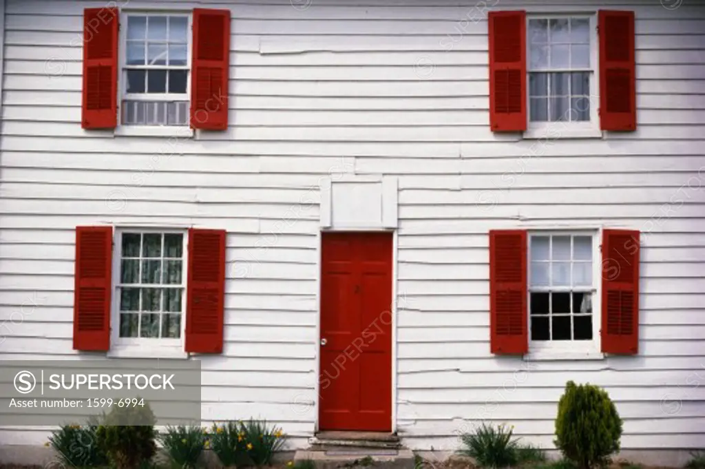 This is a close up of a white house with red shutters. The front door is also red. There are a few small bushes in front of the house.