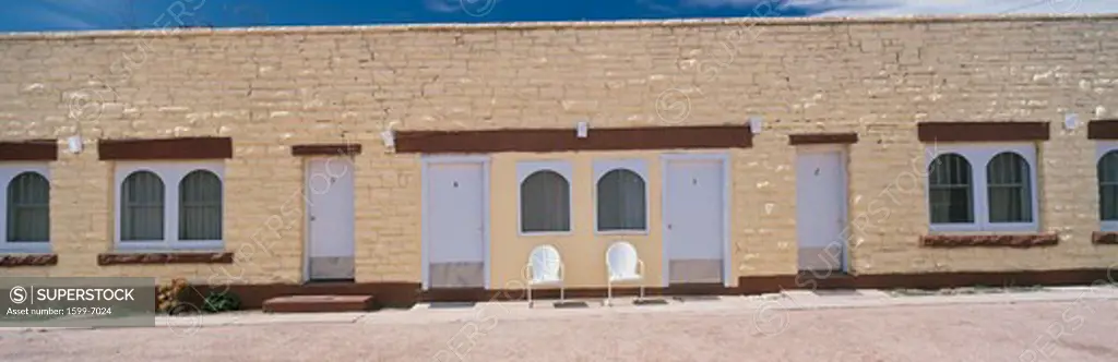This is a yellow brick motel on Route 66. it sits next to the road. There are two chairs in front of the motel.  There are windows spaced across the motel and closed doors to the hotel rooms. 