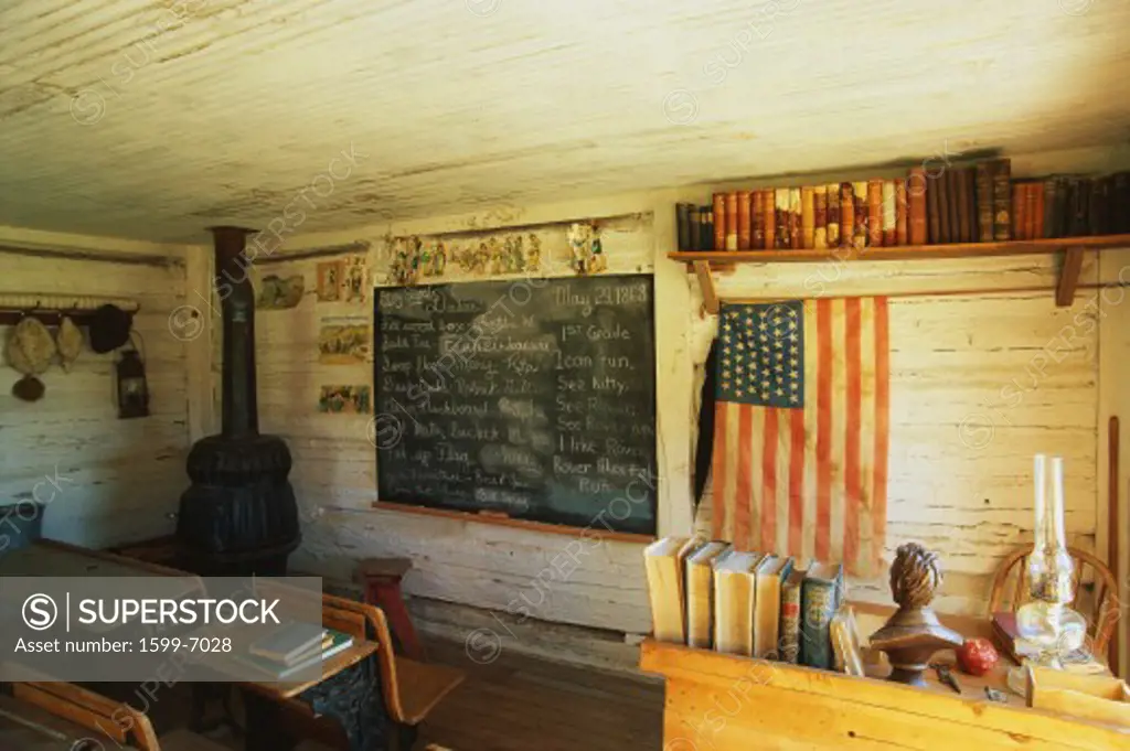 This is the interior of a one room school house. It was the first school in Montana from 1868. There is a black chalkboard and American flag hanging on the wall with a black wood furnace stove in the corner. There are a couple of old fashioned school desks to the left.