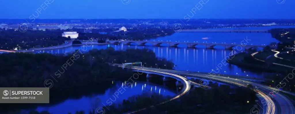 This is an aerial view of Washington, DC with the Jefferson Memorial, U.S. Capitol, Washington Monument, and Lincoln Memorial. The Potomac River runs through the center with the Key Bridge at right . There is green foliage of the spring with the streaked lights of the freeway in the foreground.