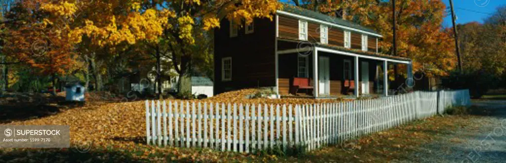 This is a brown, wooden house in a historic village near the Delaware River. A white picket fence surrounds the house. There are autumn leaves on the trees surrounding the property.
