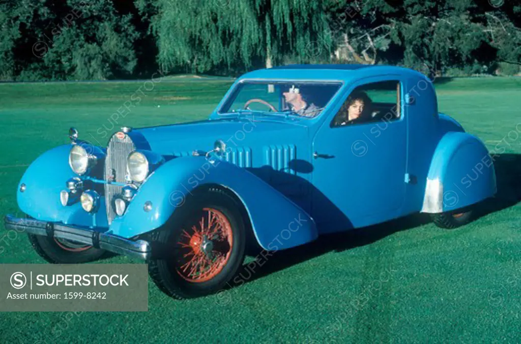 A man and woman sit in a blue Bugatti automobile at a vintage car show in Pebble Beach, California, ca. 1985.