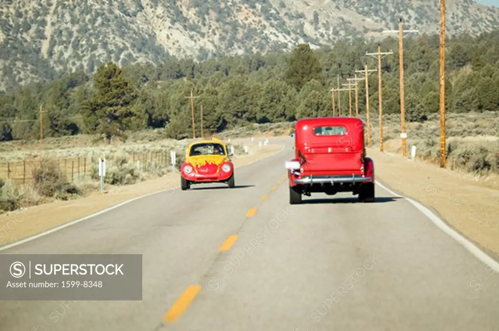 A yellow and red VW hotrod drives in opposite direction of a restored bright Red Roadster hotrod pickup truck along rural highway in Kern County near Lockwood Valley, CA