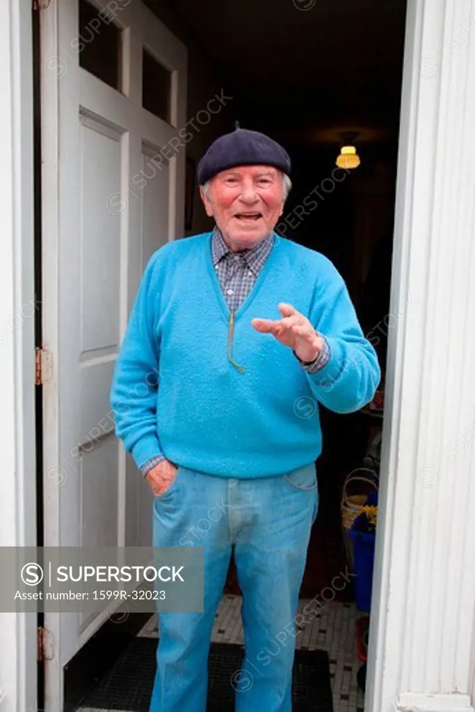Senior Italian-American man in blue seater and matching hat gestures and smiles in front of his home in the historic North End, Italian section of Boston, MA