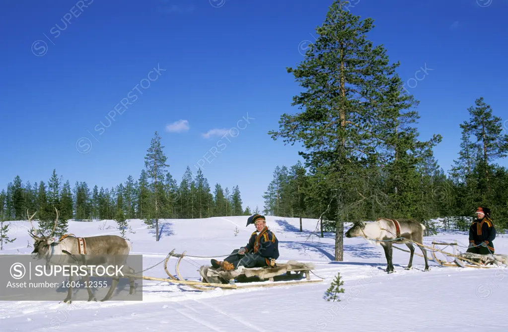 Finland, Lapland, Rovaniemi, two men in traditional costume sitting on sleights dragged by reindeers, snowy landscape